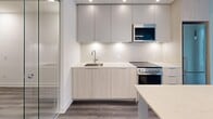 Located in Toronto, Ontario, this modern one-bedroom, two-bathroom unit offers a thoughtfully designed layout across a single floor. The contemporary kitchen is a standout feature, equipped with light wood flat-panel cabinetry, upper grey cabinets, a stone countertop with an undermount sink, stainless steel appliances including a range and built-in microwave, and under-cabinet lighting that adds a warm, polished ambiance. Dark hardwood-style flooring flows through the main living areas, grounding the bright, neutral palette of white and soft grey tones throughout. The bedroom is a comfortable, well-proportioned room featuring a large window that frames views of the surrounding urban Toronto architecture, allowing natural light to fill the space. The unit includes two bathrooms: one features a clean, glass-enclosed walk-in shower alongside a floating light wood vanity with a mirrored medicine cabinet and large-format white tile flooring, while the second includes a soaking tub set against floor-to-ceiling cream tiles with sleek chrome fixtures. Two hallways provide connectivity between spaces, and a dedicated closet adds practical storage. The overall design is clean and minimalist, reflecting a modern urban aesthetic well-suited to contemporary city living.