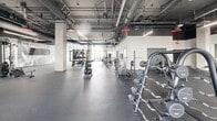 Located at 915 Division St in Nashville's Midtown neighborhood, this large-scale fitness center offers a comprehensive exercise environment spread across a single, expansive floor. The space showcases an industrial-modern aesthetic, with exposed ductwork and structural concrete columns rising to high ceilings, complemented by globe pendant lighting and bold red accents that add visual energy throughout.

The facility is divided into distinct functional zones. A dedicated studio room features light hardwood flooring, floor-to-ceiling glass walls, and ample space for yoga, stretching, and group fitness classes, with yoga mats and equipment racks at the ready. Large windows throughout the studio and main floor flood the space with abundant natural light and offer views of the surrounding Nashville skyline.

The main fitness floor is outfitted with an extensive array of Matrix strength training machines, free weights, barbells, and benches, supported by bold graphic wall murals that energize the environment. Full-length mirrors line multiple walls, enhancing the sense of openness. A dedicated cardio zone along the windowed perimeter houses treadmills, stationary bikes, and elliptical machines, each positioned to take advantage of the panoramic city views. Rubber flooring throughout the training areas ensures both safety and durability, making this a well-appointed, professional-grade fitness facility.