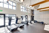 Located along Coast Highway in Dana Point, California, this fitness center delivers a well-appointed workout environment designed with both functionality and aesthetic appeal in mind. The space is defined by dramatically high ceilings that amplify the sense of openness throughout, while an extensive band of clerestory windows along the upper walls floods the interior with generous natural light and offers views of the surrounding coastal community.

The cardio zone is equipped with multiple treadmills, elliptical trainers, stationary bikes, and rowing machines, all positioned to take advantage of the bright, airy atmosphere. Large glass doors and windows at floor level further connect the space to the outdoors. A dedicated strength training area features a comprehensive free-weight section with a full dumbbell rack, adjustable benches, and large wood-framed mirrors that visually expand the room and assist with form. Cable machines and a functional training rig provide additional versatility.

Textured concrete accent walls introduce an industrial character that is balanced by warm wood beam details and nautical-themed mural artwork, nodding to the area's coastal identity. Pendant lighting and ceiling fans are suspended throughout, ensuring comfort during workouts. The durable rubber flooring provides a practical, slip-resistant surface appropriate for a full range of fitness activities, completing this well-rounded facility.