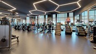 This single-floor fitness center is designed with a sophisticated, contemporary aesthetic that balances functionality with refined architectural detail. The space is defined by a striking hexagonal LED lighting system mounted against a dark ceiling, casting bright, even illumination throughout the main exercise areas and creating a visually dynamic overhead feature.

The cardio zone is generously equipped with rows of treadmills, ellipticals, stair climbers, stationary bikes, and rowing machines, all positioned to take advantage of floor-to-ceiling windows fitted with roller shades. These expansive windows admit ample natural light and offer views of the surrounding urban environment. Durable rubber flooring spans this area, providing both traction and impact absorption.

The strength training section features a comprehensive selection of free weights, weight racks, benches, cable machines, and specialty equipment. Large wall-mounted mirrors enhance spatial depth and allow for form monitoring. A dedicated entry corridor with warm wood-toned wall paneling and light hardwood-style flooring connects the zones, with a welcoming bench providing a transitional area.

A separate studio room offers a quieter setting for stretching or individual training, featuring a large framed mirror, natural light from street-facing windows, and clean, neutral finishes throughout.