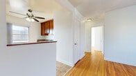 This two-bedroom, one-bathroom unit offers an efficient single-floor layout designed to maximize every square foot. Warm hardwood floors run throughout the main living areas and bedrooms, lending a cohesive and inviting character to the space. The living area is anchored by a ceiling fan and connects seamlessly to the kitchen, which features rich wood cabinetry, granite-look countertops, a stainless steel dishwasher, and tile flooring. A breakfast bar-style partition subtly separates the kitchen from the living space while maintaining an open feel. Natural light enters through multiple windows, each fitted with a radiator beneath, providing efficient heating throughout the unit. The primary bedroom offers a comfortable footprint with a sliding-door closet for storage, while the second bedroom mirrors the same hardwood flooring and bright, neutral walls. The bathroom includes a full soaking tub with white tile surround, classic mosaic tile flooring in warm terracotta tones, and a window for ventilation. A hallway connects all rooms, contributing to a functional and straightforward floor plan.