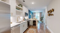 This thoughtfully designed single-floor residence seamlessly blends modern aesthetics with practical functionality. The open-concept layout connects the kitchen and living areas, creating a bright and inviting atmosphere. The kitchen is a standout feature, outfitted with white shaker-style cabinetry adorned with brushed gold hardware, sleek quartz countertops, a chevron-patterned tile backsplash, and a full suite of stainless steel appliances including a gas range and built-in microwave. Open shelving adds a decorative touch, while a built-in wine cooler at the peninsula provides a refined transition into the living space. The living area is bathed in natural light from large windows and offers direct access to a private balcony with outdoor views. A bold teal accent wall adds a vibrant focal point to this shared space. The bedroom is a serene retreat, featuring plush carpeting, generous natural light from large windows, and a rich forest-green accent wall complemented by decorative shelving. A door leads directly into the en-suite bathroom, which is finished with large-format tile flooring, a white vanity with gold hardware, and a glass-enclosed walk-in shower. Conveniently accessible from the bathroom, a dedicated laundry area houses a stacked full-size washer and dryer alongside built-in wire shelving for additional storage. Warm hardwood-style flooring flows throughout the main living areas, tying the residence together with a cohesive and polished finish.