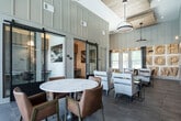 Located in Huntsville, Alabama, this single-story community clubhouse showcases a sophisticated blend of modern farmhouse and contemporary design. The space is defined by soaring ceilings with warm wood-paneled accents and vertical shiplap walls finished in soft neutral tones, creating an atmosphere that feels both refined and welcoming.

The main lounge area is anchored by plush light gray seating arrangements paired with a dark metal coffee table, set beneath a striking circular pendant light. A bold decorative wall installation featuring oversized carved wooden letters adds an artistic focal point, complementing the curated framed artwork displayed throughout. A round marble-topped dining table surrounded by rich cognac leather chairs serves as a stylish gathering spot.

Large floor-to-ceiling windows flood the space with natural light and offer views of the surrounding outdoor areas. Sliding barn-style doors with black metal hardware and glass panels lead to private rooms suitable for work or meetings, with one area visible as a dedicated workspace.

The flooring transitions between large-format dark tile in the lounge and a geometric patterned wood-look tile in the connecting corridor, adding visual interest and defining distinct zones within the open layout. The overall ambiance is polished yet approachable, making this clubhouse an inviting hub for community life.