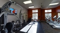 This fitness center is a dedicated single-floor exercise facility designed to accommodate a full range of workout routines. The space features a bold accent wall in deep orange, contrasted by neutral gray walls, creating an energizing and motivating atmosphere. High ceilings amplify the sense of openness, while large windows along the exterior wall flood the room with natural light, offering views of the outdoors and enhancing the overall workout environment. A ceiling fan aids in air circulation throughout the space. The facility is outfitted with a multi-station cable machine for strength training, a dedicated free weight area complete with a tiered dumbbell rack and an adjustable weight bench, and cardio equipment including a treadmill and additional exercise machines. Rubber flooring provides a safe and durable surface across the workout area, transitioning to hard flooring near the perimeter. A wall-mounted flat-screen television is positioned for visibility from multiple workout stations. Recessed and flush-mounted ceiling lights supplement the natural light, ensuring the space is well-lit at all times. This fitness center is thoughtfully arranged to maximize usability and comfort for users of all fitness levels.