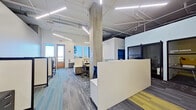 This contemporary single-floor office space combines industrial architectural elements with a clean, functional design to create a productive and visually engaging work environment. Exposed concrete columns and an open ceiling with visible ductwork, piping, and linear LED lighting fixtures give the space a modern industrial character, while white walls and patterned carpet tiles in shades of blue, gray, and yellow provide a polished, professional finish.

The open-plan work area is thoughtfully organized with multiple workstation clusters, each featuring paneled privacy dividers, ergonomic task chairs, and built-in storage pedestals with a wood-grain finish. A prominent white reception or divider counter anchors the central area, clearly delineating zones within the open floor plan.

Along the perimeter, large windows flood the interior with natural light and offer views of the surrounding exterior, contributing to an airy, open atmosphere. A glazed private office or meeting room enclosed with dark-framed barn-style sliding doors adds a touch of refined contrast and provides a semi-private workspace option.

A casual lounge nook with a built-in bench seat, accent lighting, and a decorative feature wall offers a relaxed alternative to traditional desk seating. The overall layout balances collaborative open-plan working with privacy, making it well-suited for a professional office or coworking environment.