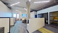 This contemporary single-floor office space combines industrial architectural elements with a clean, functional design to create a productive and visually engaging work environment. Exposed concrete columns and an open ceiling with visible ductwork, piping, and linear LED lighting fixtures give the space a modern industrial character, while white walls and patterned carpet tiles in shades of blue, gray, and yellow provide a polished, professional finish.

The open-plan work area is thoughtfully organized with multiple workstation clusters, each featuring paneled privacy dividers, ergonomic task chairs, and built-in storage pedestals with a wood-grain finish. A prominent white reception or divider counter anchors the central area, clearly delineating zones within the open floor plan.

Along the perimeter, large windows flood the interior with natural light and offer views of the surrounding exterior, contributing to an airy, open atmosphere. A glazed private office or meeting room enclosed with dark-framed barn-style sliding doors adds a touch of refined contrast and provides a semi-private workspace option.

A casual lounge nook with a built-in bench seat, accent lighting, and a decorative feature wall offers a relaxed alternative to traditional desk seating. The overall layout balances collaborative open-plan working with privacy, making it well-suited for a professional office or coworking environment.
