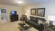 This single-floor space features a thoughtfully arranged living area that balances comfort and style. The living room serves as the focal point, showcasing neutral beige walls that create a warm, welcoming ambiance. Plush, light-toned carpeting runs underfoot, adding a soft texture that enhances the room's cozy feel. A large, charcoal-grey sofa anchors the seating area, complemented by a classic striped accent chair that adds visual interest. A sleek, glass-topped coffee table sits atop a shaggy area rug, providing both function and a layered sense of design. Dark wood furniture, including a multi-drawer media console, grounds the space and provides ample storage. A flat-screen television is positioned for comfortable viewing. The walls are adorned with framed nature-inspired artwork featuring bird motifs, adding a refined decorative touch. Ambient lighting from a central ceiling fixture and a table lamp creates a soft, even glow throughout the room. The textured ceiling adds subtle architectural character, while the overall layout makes efficient use of the available space, resulting in a functional and inviting living environment.