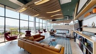 This expansive rooftop amenity lounge is a showcase of bold contemporary design, offering an elevated communal experience with sweeping panoramic views of the surrounding cityscape and greenery through dramatic floor-to-ceiling glass walls. The space's most distinctive architectural feature is its undulating wooden wave ceiling installation, which ripples organically across the room and is accented by integrated lighting that casts a warm, ambient glow throughout.

The main lounge area is thoughtfully furnished with a mix of seating arrangements, including rich leather sectional sofas, modern accent chairs in deep burgundy and earthy tones, and intimate conversation groupings centered on patterned area rugs in teal and blue. A full-width linear gas fireplace set into a light stone surround serves as a striking focal point, flanked by additional lounge seating for a cozy yet sophisticated atmosphere.

A dedicated entertainment and media wall features bold geometric wood and dark panel detailing alongside a mounted flat-screen display. The space also includes a fully equipped kitchen with light wood cabinetry, a mosaic tile backsplash in soft aqua tones, stainless steel appliances, and a generous marble-topped island with bar seating — ideal for hosting and social gatherings. Light hardwood flooring flows throughout, and access to an outdoor terrace further extends the space's appeal.