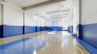 This indoor basketball court is a well-appointed athletic facility designed to support a wide range of sports and recreational activities. The space is defined by its soaring ceilings, which provide generous vertical clearance well-suited for competitive and casual play alike. The polished hardwood floor is finished with crisp blue and white court markings, including boundary lines, a center circle, and key areas, ensuring a professional-grade playing surface. Blue padded wall panels line the lower perimeter of the court, offering both a safety buffer for players and a clean, cohesive aesthetic. A regulation-height basketball backboard and hoop are mounted at one end of the court, with the opposing end offering clear sightlines across the full length of the floor. Natural light filters in through large multi-pane windows positioned high along one side wall, complemented by industrial pendant lighting suspended from the coffered ceiling structure above. The white-painted upper walls and ceiling help reflect light evenly throughout the space, maintaining a bright and energetic atmosphere. A bench along one wall provides seating for spectators or resting players. The overall design balances functionality with a clean, institutional aesthetic, making it a versatile venue for basketball, fitness programs, and group activities.