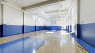 This indoor basketball court is a well-appointed athletic facility designed to support a wide range of sports and recreational activities. The space is defined by its soaring ceilings, which provide generous vertical clearance well-suited for competitive and casual play alike. The polished hardwood floor is finished with crisp blue and white court markings, including boundary lines, a center circle, and key areas, ensuring a professional-grade playing surface. Blue padded wall panels line the lower perimeter of the court, offering both a safety buffer for players and a clean, cohesive aesthetic. A regulation-height basketball backboard and hoop are mounted at one end of the court, with the opposing end offering clear sightlines across the full length of the floor. Natural light filters in through large multi-pane windows positioned high along one side wall, complemented by industrial pendant lighting suspended from the coffered ceiling structure above. The white-painted upper walls and ceiling help reflect light evenly throughout the space, maintaining a bright and energetic atmosphere. A bench along one wall provides seating for spectators or resting players. The overall design balances functionality with a clean, institutional aesthetic, making it a versatile venue for basketball, fitness programs, and group activities.