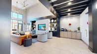 Located at 2611 Hopps Rd in Elgin, IL, this contemporary community amenity space is thoughtfully designed to deliver both comfort and functionality across a single, well-appointed floor. The grand entrance hallway welcomes visitors with soaring ceilings, bold dark structural beams, and large-format light tile flooring that flows throughout the space.

The central lounge area serves as the heart of the facility, anchored by a striking white stacked-stone feature wall with a built-in flat-screen display and flanked by sleek, dark shelving with decorative accents. Curved lounge seating and a warm mix of modern furnishings create an inviting social atmosphere. A beverage and refreshment station with subway tile backsplash sits conveniently nearby.

Floor-to-ceiling glass walls and expansive sliding doors flood the space with natural light and open directly onto a furnished outdoor patio, complete with a pergola, dining seating, and views of the surrounding pool area—seamlessly blending indoor and outdoor living.

The full kitchen is outfitted with light wood cabinetry, stainless steel appliances, a white quartz-topped island with bar seating, and a subway tile backsplash, making it ideal for gatherings and events.

The fitness center features blue accent walls, full-length mirrors, rubber flooring, ceiling fans, and a comprehensive array of strength and cardio equipment, including cable machines, free weights, and benches—offering a complete workout experience with natural light from large windows.
