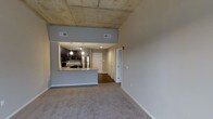 This single-floor unit showcases a modern industrial aesthetic defined by its striking exposed concrete ceiling, which adds texture and an urban character to the open-concept layout. The living area is finished with neutral wall-to-wall carpeting, creating a soft contrast against the raw overhead surface and contributing to a comfortable, livable atmosphere.

The kitchen is positioned prominently along one wall and is separated from the living space by a raised granite-topped peninsula, which provides both a functional workspace and casual dining or entertaining opportunities. Dark cabinetry lines the kitchen walls, complemented by stainless steel appliances including a refrigerator, oven range, and microwave. Pendant lighting above the counter adds warmth and visual definition to the cooking area.

A hallway extends from the main living space toward the back of the unit, where multiple doors suggest access to closets or additional rooms. The flooring transitions from carpet in the living area to hard flooring in the hallway, adding a practical distinction between zones. The neutral gray walls throughout contribute to a cohesive and contemporary overall palette.