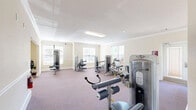 Located at 2006 Karl Dr in Warner Robins, Georgia, this dedicated fitness facility offers a generously sized, single-floor exercise space designed to support a comprehensive workout experience. The interior is finished with warm cream-toned walls, elegant crown molding, and wide white baseboards, lending the space a polished and well-maintained aesthetic. Plush mauve carpeting covers the floors throughout, providing both comfort and durability underfoot.

The facility is organized into distinct workout zones. A cardio area features multiple treadmills and elliptical trainers positioned near large windows fitted with white blinds, allowing natural light to fill the room. A ceiling fan overhead ensures comfortable air circulation during workouts. The strength training zone is well-stocked with a variety of selectorized cable and resistance machines, catering to a full range of muscle groups. Additional strength equipment is distributed throughout the space, offering users ample room to move freely between stations.

Overhead fluorescent lighting supplements the natural light from multiple windows, keeping the space bright and functional at any time of day. A fire extinguisher is mounted on the wall, reflecting attention to safety. The thoughtful layout, quality finishes, and broad equipment selection make this fitness center a practical and inviting facility for residents seeking a convenient place to maintain an active lifestyle.