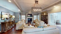 This single-story clubhouse in Daphne, Alabama, offers a well-appointed gathering space designed with a coastal aesthetic throughout. The main lounge area serves as the centerpiece, featuring rich hardwood floors, crisp crown molding, and a tray ceiling accented by a striking rope-and-metal chandelier. A white-mantled fireplace with a wall-mounted television above anchors the room, flanked by plantation-shuttered windows that allow ample natural light. Comfortable striped upholstered seating and wicker accent chairs are arranged around a substantial wooden coffee table, creating an inviting atmosphere for socializing.

The walls combine soft gray and coastal blue tones, divided by a chair rail that adds architectural character. Built-in bookshelves along one wall provide both storage and visual interest. A dedicated workspace with a wooden desk, chair, and monitor offers a functional area for administrative use. French doors connect the lounge to an adjacent room, extending the usable space.

The kitchenette features dark cabinetry with granite countertops, stainless steel appliances including a full-size refrigerator and microwave, and a pass-through counter opening to the lounge area. Throughout, recessed lighting and decorative lamps ensure well-lit, welcoming interiors suited for community gatherings and events.
