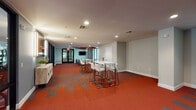This single-floor amenity center is designed with a cohesive, energetic aesthetic anchored by a striking red and gray patterned carpet that flows throughout the space. The fitness area is generously sized and bathed in natural light through floor-to-ceiling windows that overlook an outdoor pool and surrounding landscaping. It is equipped with a comprehensive selection of Life Fitness cardio machines, including treadmills, ellipticals, and stationary bikes, as well as a multi-functional training rig with a punching bag, TRX suspension trainers, and free weight benches. A bold rock-climbing mural decorates one wall, adding visual energy to the workout environment. Adjacent to the gym floor, a built-in counter with dark wood cabinetry, a light stone countertop, and an under-counter beverage refrigerator provides a convenient refreshment station. The lounge area features a distinctive circular wood-paneled ceiling medallion above a seating arrangement of upholstered armchairs centered around a round coffee table, with a wall-mounted television and sheer curtain-framed windows. A separate gathering area offers bar-height white tables with white upholstered stools for social or collaborative use. Large glass doors provide direct access to the outdoor pool courtyard, connecting the interior seamlessly to the exterior amenities.