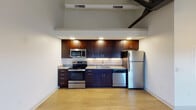 This single-floor unit showcases a loft-inspired design, characterized by dramatically high ceilings with exposed dark steel beams and industrial-style ceiling fans that add architectural interest throughout the space. The open layout is anchored by a well-appointed kitchen featuring rich, dark-toned cabinetry, granite-look countertops with a subway tile backsplash, and a full suite of stainless steel appliances including a range, microwave, dishwasher, and refrigerator. Under-cabinet lighting enhances both functionality and ambiance in the kitchen area.

Light hardwood flooring runs throughout the unit, providing warmth and continuity across each room. The main living area benefits from soaring vaulted ceilings and multiple windows that invite generous natural light, offering views to the surrounding exterior. Two interior doors off the main living space lead to additional rooms, each featuring their own windows with natural light.

The bathroom is finished with classic penny-tile flooring, a dark vanity with a white countertop, and clean white walls, creating a crisp, timeless aesthetic. The overall design effectively balances industrial architectural elements with modern, functional finishes, resulting in a bright and distinctive living space.