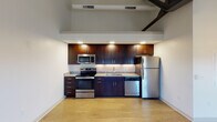 This single-floor unit showcases a loft-inspired design, characterized by dramatically high ceilings with exposed dark steel beams and industrial-style ceiling fans that add architectural interest throughout the space. The open layout is anchored by a well-appointed kitchen featuring rich, dark-toned cabinetry, granite-look countertops with a subway tile backsplash, and a full suite of stainless steel appliances including a range, microwave, dishwasher, and refrigerator. Under-cabinet lighting enhances both functionality and ambiance in the kitchen area.

Light hardwood flooring runs throughout the unit, providing warmth and continuity across each room. The main living area benefits from soaring vaulted ceilings and multiple windows that invite generous natural light, offering views to the surrounding exterior. Two interior doors off the main living space lead to additional rooms, each featuring their own windows with natural light.

The bathroom is finished with classic penny-tile flooring, a dark vanity with a white countertop, and clean white walls, creating a crisp, timeless aesthetic. The overall design effectively balances industrial architectural elements with modern, functional finishes, resulting in a bright and distinctive living space.