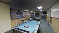This single-floor recreational facility is designed as a social and entertainment venue. The main area is anchored by two full-sized pool tables with blue felt surfaces, positioned prominently throughout the open floor plan. A built-in bar counter with wood panel detailing runs along one side of the room, accompanied by bar stools, suggesting a service or refreshment area. The ceiling features a suspended tile grid with recessed fluorescent panel lighting that evenly illuminates the space. Wall-mounted televisions are positioned for easy viewing throughout the room. Wood paneling lines the lower portion of the walls, contributing a utilitarian yet classic aesthetic. Blue-framed doorways lead to additional areas, and a map is displayed on one wall, adding a functional decorative element. A seating area with casual furniture is tucked toward the back, offering a relaxed spot away from the game tables. The overall ambiance is casual and functional, characteristic of a club room, community lounge, or recreational hall suited for group activities and social interaction.