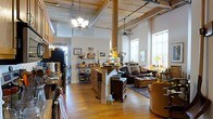 This single-floor loft-style space captivates with its dramatic architectural character and open, airy layout. Soaring ceilings clad in natural wood planks and supported by exposed structural beams create a warm, industrial-rustic ambiance throughout. Tall windows along one wall flood the interior with abundant natural light, enhancing the sense of spaciousness.

The open floor plan connects the kitchen, living, and workspace areas in a fluid, continuous layout. The kitchen features warm wood cabinetry, a freestanding island with built-in storage, and an overhead chandelier that adds a classic touch. Track lighting runs along the ceiling beams, offering practical illumination throughout the space.

A central structural column anchors the room visually while allowing the living area to flow naturally from the kitchen. The living space is appointed with rich, dark leather sofas arranged around a traditional area rug, providing a cozy contrast to the loft's industrial bones. Wood furnishings and layered décor add depth and personality.

A ceiling fan ensures comfortable airflow, while the overall combination of reclaimed-style wood ceilings, hardwood floors, exposed columns, and generous natural light gives this loft a distinctive, character-filled quality that seamlessly blends rustic warmth with open, modern living.