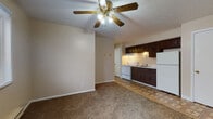 This single-floor unit offers a practical and straightforward layout designed for comfortable everyday living. The living area is finished with wall-to-wall carpeting and neutral beige walls, providing a warm, understated atmosphere. A ceiling fan with integrated lighting serves as a central fixture, while a window unit air conditioner and baseboard heating ensure year-round climate control. Windows allow natural light to filter in, offering views of the surrounding outdoors.

The kitchen is positioned adjacent to the living space and features dark wood cabinetry — both upper and lower — paired with light countertops, a double-basin stainless steel sink, a white electric coil range, and a white refrigerator. A range hood and under-cabinet lighting provide functional illumination for meal preparation. The kitchen floor is covered in a patterned vinyl tile, distinguishing it from the carpeted living areas.

The bedroom is a generously sized room with plush carpeting, a ceiling fan, a window with blinds for natural light and privacy, and a built-in closet with louvered bifold doors offering ample storage.

The bathroom is clean and efficient, featuring a white tiled tub and shower surround, a toilet, a single-basin vanity with a marbled countertop, and a mirrored medicine cabinet above. The floor is finished in a stone-look tile.
