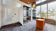 Located at 337 NE 103rd St in Seattle, WA, this contemporary single-floor workspace delivers a polished, functional environment designed for focused work and collaboration. The space makes a strong visual impression with its soaring ceilings, slatted wood ceiling panels with recessed lighting, and exposed structural elements that lend an industrial-modern character.

A defining feature of the layout is the inclusion of freestanding glass acoustic pods — self-contained work booths equipped with interior seating, task lighting, and soundproofing for private calls or focused sessions. These pods are complemented by an open lounge area furnished with rounded, orange upholstered chairs and a compact table, ideal for informal meetings or collaborative work.

Biophilic design is evident throughout, most notably in a framed moss-and-greenery wall art piece that adds a vibrant, organic accent to the otherwise neutral palette of white walls and patterned gray carpet tiles. A sleek teal credenza beneath the artwork adds a refined, modern touch.

Large windows along one wall usher in generous natural light and offer views of the surrounding Seattle streetscape and tree canopy, enhancing the sense of openness despite the intimate scale. The overall ambiance balances professionalism with warmth, making this a versatile and inviting workspace.