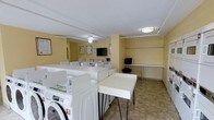 This communal laundry facility is designed with both functionality and comfort in mind. The room is finished with warm yellow walls and neutral tile flooring, creating a bright and welcoming atmosphere. Overhead, flush-mount and ceiling fan light fixtures provide ample illumination throughout the space.

The room is outfitted with a generous number of front-loading and top-loading washers and dryers, arranged in organized rows to accommodate multiple users simultaneously. Spacious white folding tables are positioned conveniently throughout the room, offering plenty of surface area for sorting and folding laundry. A dedicated shelving unit along one wall provides additional storage or staging space, complemented by a wall-mounted clothing rod ideal for hanging garments.

A wall-mounted flat-screen television is positioned for easy viewing, allowing users to stay entertained during their visit. Informational notices and posted guidelines are displayed on the walls, keeping users informed about facility rules and procedures. The layout is open and thoughtfully arranged, ensuring easy circulation between machines and work surfaces. Overall, this laundry facility offers a clean, practical, and well-maintained environment suited to serving a shared residential community.