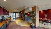 This single-floor business center in Houston, TX offers a thoughtfully designed commercial space with a distinctive and energetic aesthetic. The interior is characterized by a warm, bold color palette — deep crimson accent walls contrast with earthy brown tones and light hardwood flooring that flows through the main area, while patterned carpet zones define separate functional spaces.

Upon entry through the glass-paneled double doors, visitors are welcomed into an open reception area anchored by a striking checkerboard-tiled bar counter with bar-height seating. A tall, lush indoor tree adds a natural element near the entrance. Track lighting runs along a curved ceiling detail, guiding the eye through the space and illuminating key areas.

A dedicated refreshment station lines one wall, equipped with dark cabinetry, a mini-refrigerator, countertop appliances, and ample counter space. Adjacent to this, a rustic wood communal table set against a bold red wall provides a casual meeting or dining area, complemented by framed wall art.

A lounge area features comfortable tan leather armchairs arranged around a gold accent table, positioned beside expansive floor-to-ceiling windows that draw in natural light and offer exterior views. A workstation area with multiple desks and computers rounds out the functional offerings of this well-appointed space.