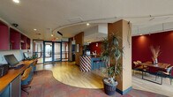 This single-floor business center in Houston, TX offers a thoughtfully designed commercial space with a distinctive and energetic aesthetic. The interior is characterized by a warm, bold color palette — deep crimson accent walls contrast with earthy brown tones and light hardwood flooring that flows through the main area, while patterned carpet zones define separate functional spaces.

Upon entry through the glass-paneled double doors, visitors are welcomed into an open reception area anchored by a striking checkerboard-tiled bar counter with bar-height seating. A tall, lush indoor tree adds a natural element near the entrance. Track lighting runs along a curved ceiling detail, guiding the eye through the space and illuminating key areas.

A dedicated refreshment station lines one wall, equipped with dark cabinetry, a mini-refrigerator, countertop appliances, and ample counter space. Adjacent to this, a rustic wood communal table set against a bold red wall provides a casual meeting or dining area, complemented by framed wall art.

A lounge area features comfortable tan leather armchairs arranged around a gold accent table, positioned beside expansive floor-to-ceiling windows that draw in natural light and offer exterior views. A workstation area with multiple desks and computers rounds out the functional offerings of this well-appointed space.