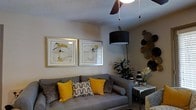 This single-floor space showcases a well-appointed living area designed with a contemporary aesthetic and a carefully coordinated color scheme. The room centers around a plush grey sofa adorned with an eye-catching mix of throw pillows in mustard yellow, black-and-white geometric patterns, and textured grey tones, creating a layered and inviting seating arrangement.

Overhead, a combination of a ceiling fan and a bold black drum pendant light provides both functional and ambient lighting, contributing to the room's modern character. The walls are painted in a warm neutral tone, serving as a clean backdrop for two matching framed art pieces featuring abstract floral motifs with yellow and black accents, displayed side by side for a gallery-like effect.

A small marble-top side table sits adjacent to the sofa, styled with decorative objects and fresh white florals, adding an elegant touch. Lush indoor greenery introduces a natural element, while a collection of metallic disc wall décor on the opposite wall adds visual texture and depth. The overall ambiance is sophisticated yet comfortable, with consistent attention to detail throughout.
