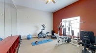 This home gym is designed to support a well-rounded fitness routine within a comfortable, dedicated space. The room features a striking deep red accent wall that adds energy and visual contrast to the otherwise neutral, light-toned walls, creating a motivating atmosphere suited for exercise. Carpet flooring runs throughout, providing cushioning underfoot, while a ceiling fan overhead ensures adequate airflow during workouts.

Natural light enters through a window fitted with blinds on the right side of the room, keeping the space bright without harsh glare. The room accommodates a multi-station cable and weight machine, ideal for strength training, positioned near the window. An elliptical cross-trainer occupies the center of the room, paired with a blue exercise mat and a set of free weights for floor-based workouts. A stability ball adds further versatility for core training exercises.

The layout is thoughtfully arranged to maximize usable floor space while keeping equipment accessible. The ceiling fan and window ventilation contribute to a practical, comfortable environment, making this room a well-equipped and functional space for regular home fitness use.