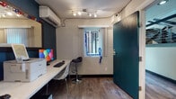 This single-floor workspace is thoughtfully designed to maximize functionality within a compact footprint. The main area centers around a long, white marble-topped built-in desk that spans one wall, providing ample surface area for computing and office tasks. The workstation is equipped with a desktop computer and printer, making it ready for immediate use. Seating options include a sleek black bar-height stool and a modern lattice-style chair, offering flexibility for different work styles.

The space is defined by a striking deep teal accent wall that adds a bold, contemporary character, contrasting with the neutral beige textured walls throughout. Wood-look plank flooring runs across the entire floor, lending warmth and visual continuity. A wall-mounted split air conditioning unit ensures climate comfort year-round.

Overhead, a multi-head track lighting fixture provides directed, adjustable illumination across the workspace. A single window with vertical blinds allows natural light to filter in. A large, vibrant abstract canvas painting adorns one of the neutral walls, adding an artistic focal point to the space. The teal-painted entry door reinforces the cohesive color palette. A security camera is mounted near the ceiling, reflecting attention to safety and security within this well-appointed, practical workspace.
