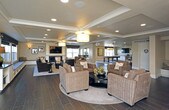 This single-floor community clubhouse is designed to offer residents a versatile and comfortable gathering space. The entrance lobby sets a welcoming tone with rich dark hardwood-style flooring, recessed lighting, decorative wall art, and an open seating arrangement featuring upholstered armchairs around a central coffee table, accented by lush indoor plants that bring a touch of nature indoors.

The main lounge area is a standout feature of the space, showcasing a coffered ceiling with a central drum pendant light and recessed fixtures. Built-in window bench seating lines the perimeter, offering views of the outdoor playground area. A central fireplace framed by a clean white surround serves as the focal point, with a wall-mounted flat-screen television above it. Plush brown sofas and wicker accent chairs are arranged around a patterned area rug, creating an inviting atmosphere for socializing or relaxing.

Adjacent to the lounge is a spacious game room anchored by a full-size billiards table set atop a neutral area rug on expansive dark hardwood floors. A textured stone accent wall adds visual depth behind a mounted television, while a built-in wet bar with bar stool seating provides a dedicated entertaining hub. Pendant lighting and recessed fixtures ensure the space is well-lit throughout, making this clubhouse a comprehensive amenity for community gatherings.