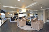 This single-floor community clubhouse is designed to offer residents a versatile and comfortable gathering space. The entrance lobby sets a welcoming tone with rich dark hardwood-style flooring, recessed lighting, decorative wall art, and an open seating arrangement featuring upholstered armchairs around a central coffee table, accented by lush indoor plants that bring a touch of nature indoors.

The main lounge area is a standout feature of the space, showcasing a coffered ceiling with a central drum pendant light and recessed fixtures. Built-in window bench seating lines the perimeter, offering views of the outdoor playground area. A central fireplace framed by a clean white surround serves as the focal point, with a wall-mounted flat-screen television above it. Plush brown sofas and wicker accent chairs are arranged around a patterned area rug, creating an inviting atmosphere for socializing or relaxing.

Adjacent to the lounge is a spacious game room anchored by a full-size billiards table set atop a neutral area rug on expansive dark hardwood floors. A textured stone accent wall adds visual depth behind a mounted television, while a built-in wet bar with bar stool seating provides a dedicated entertaining hub. Pendant lighting and recessed fixtures ensure the space is well-lit throughout, making this clubhouse a comprehensive amenity for community gatherings.