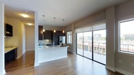 This contemporary single-floor residence showcases a thoughtfully designed open-concept layout that seamlessly blends the kitchen and living areas into one cohesive, light-filled space. The kitchen is a standout feature, outfitted with dark espresso cabinetry that creates a striking contrast against the crisp white countertops and light hardwood flooring. A generous kitchen island anchors the space and provides additional prep and seating options. Three elegant pendant lights hang above, adding warmth and a touch of modern sophistication. Stainless steel appliances, including a French door refrigerator, complete the well-appointed kitchen. Large floor-to-ceiling sliding glass doors dominate one wall, flooding the interior with abundant natural light and providing direct access to a private balcony. The balcony offers impressive elevated views of the surrounding urban landscape, with city buildings and mature trees visible in the distance. Roller shades on the windows allow for flexible light and privacy control. The smooth, light-toned hardwood floors extend throughout, enhancing the airy, open feel of the space. The neutral color palette and clean architectural lines throughout give this residence a polished, move-in-ready aesthetic suited to modern urban living.