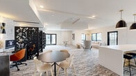 This thoughtfully designed, single-floor amenity space offers a generous and versatile layout accommodating a variety of functions. The interior is finished in a crisp, neutral palette of whites and warm beiges, accentuated by textured, patterned carpeting that runs throughout and adds visual depth underfoot.

The lounge area is a standout feature, anchored by a large sectional sofa in soft cream, a round dark coffee table, and an eye-catching gold sputnik chandelier. Large black-framed windows flood the space with natural light and offer views of lush greenery and neighboring architecture. An intimate seating nook nearby features a woven pendant light and curated abstract artwork, creating a relaxed and inviting atmosphere.

The co-working zone is defined by three individual workstations, each separated by striking black laser-cut decorative screens and illuminated by wall-mounted sconces against a textured stone accent wall. This area provides both privacy and a stylish aesthetic suited for focused work.

A dedicated conference room accommodates a full-sized wood conference table surrounded by leather executive chairs, with bold pendant lighting overhead and an artistic bird-motif wallcovering as a dramatic focal point. A mounted display screen enables presentations and meetings. French doors connect various zones, maintaining an open yet organized flow throughout the entire space.