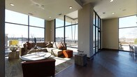 This single-floor space makes an immediate impression with its dramatic floor-to-ceiling windows that wrap around the living area, flooding the interior with abundant natural light and offering sweeping views of the surrounding cityscape. The open-plan layout creates a generous sense of space, seamlessly connecting the lounge and living areas in a cohesive, flowing design.

The living area is anchored by a substantial sectional sofa in warm earth tones, complemented by an orange accent ottoman and a understated area rug, lending the space a contemporary yet inviting character. A low-profile side table and carefully placed decorative accents — including a model airplane — add personality without cluttering the clean lines of the room.

Dark hardwood flooring runs throughout, providing a rich contrast against the light neutral walls and the bright, airy atmosphere created by the expansive glazing. Structural columns are seamlessly integrated into the design, adding architectural definition while maintaining the open feel of the space.

The steel-framed window system is a standout architectural feature, giving the space an industrial-modern edge. Recessed ceiling lighting and well-placed ventilation elements speak to a thoughtfully designed infrastructure. Overall, the space exudes a refined urban atmosphere, balancing modern design with comfort and practicality.