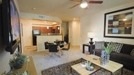 This single-floor residence offers a thoughtfully designed open-concept layout that seamlessly connects the living, dining, and kitchen areas into one cohesive and functional space. The living room is warm and inviting, anchored by a ceiling fan with integrated lighting, plush carpeting, and tasteful contemporary furnishings. A textured area rug and modern accent pieces add depth and character to the space.

The dining area sits at the heart of the floor plan, featuring a glass-top table with sleek black chairs, making it an elegant spot for everyday meals or entertaining guests. It flows naturally into the kitchen, which is separated by a raised breakfast bar and defined by rich, warm-toned cabinetry. The kitchen is well-equipped with full-size black appliances, including a refrigerator and range, along with pendant lighting and a dedicated track lighting system that illuminates the workspace effectively.

An arched architectural detail frames the transition between the kitchen and living areas, adding a refined design touch throughout. Recessed lighting and ample natural illumination contribute to the bright, airy ambiance of the entire space. The neutral color palette of warm beiges and creams throughout the walls and flooring creates a welcoming and versatile environment suitable for a variety of interior styles.