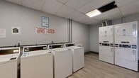 This shared laundry facility is a single-room space designed for practical, high-capacity use. The room is finished with light wood-look vinyl flooring and neutral gray walls, giving it a clean and functional aesthetic. A drop ceiling with recessed fluorescent panel lighting ensures the space is well-lit throughout.

The laundry area is equipped with a row of commercial-grade top-loading washing machines along one wall, complemented by stacked washer-dryer units along an adjacent wall, providing ample capacity for multiple users. Exposed utility pipes run along the walls, characteristic of a utility-focused shared space. Posted signage on the walls provides usage guidelines and laundry policies for users.

A folding table is available in an open area of the room, offering a convenient surface for sorting and folding laundry. The space also features a fire alarm, a security camera, and an emergency exit door, underscoring its safety-conscious design. Baseboard heating is visible along the walls, ensuring comfort during colder months. The layout is open and efficient, maximizing usability while keeping the overall footprint compact and well-organized.