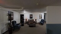 This single-floor space presents a cozy, themed lounge area with a distinct retro-cinema aesthetic. The room is furnished with a cushioned loveseat, a wingback armchair, and an additional side chair, all arranged around a wooden coffee table to create an inviting seating arrangement. Two matching table lamps on side tables provide warm, ambient lighting throughout the space. A notable decorative element is a dark sculptural statue positioned near one wall, adding a dramatic focal point to the room. The walls are adorned with framed classic movie posters, reinforcing the cinematic theme. Exposed utility pipes run along the ceiling, lending an industrial character to the otherwise traditionally styled interior. The neutral carpeted flooring and white walls keep the space feeling open and understated. A doorway is visible on the left wall, indicating access to additional areas beyond. The overall ambiance is dimly lit and intimate, evoking the nostalgic atmosphere of a classic movie theater lobby or a themed entertainment lounge.
