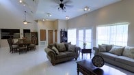 Located at 2335 Atkinson Rd in Biloxi, Mississippi, this single-story property offers a well-designed layout centered around a spacious, open living area. The room immediately impresses with its soaring vaulted ceilings, which lend an airy, expansive feel to the interior. A ceiling fan with a classic light fixture is mounted overhead, complementing the track lighting that ensures the space remains well-lit at all hours. Large French doors and wide windows line the exterior wall, inviting abundant natural light and offering views of the outdoors, creating a bright and welcoming atmosphere. The living area provides ample room for comfortable seating arrangements, as evidenced by the generous floor space and polished light-toned flooring that reflects natural light throughout. A dining area is visible to one side, complete with traditional wood furniture and a display cabinet, adding a warm, classic character to the home. Interior doors lead to additional rooms, suggesting a functional and thoughtfully organized floor plan. The overall ambiance is warm and inviting, combining practical design with comfortable living spaces suited to the Gulf Coast lifestyle.