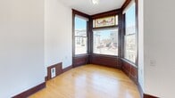 Located at 220 Iowa Ave in Muscatine, Iowa, this two-bedroom, one-bathroom unit blends historic charm with functional living across a well-organized single floor. The living area is highlighted by a striking bay window adorned with a colorful stained glass transom, framed by rich dark wood trim that adds warmth and character. Hardwood floors run through the living and common areas, complementing the bold dark baseboards and classic millwork found throughout the unit. A hallway connects the spaces efficiently, lined with beautifully crafted dark wood panel doors that reinforce the historic aesthetic. The primary bedroom is generously sized, featuring carpet flooring, a ceiling fan, and bay windows that invite natural light and street views. The second bedroom also offers carpet, a ceiling fan, and a window overlooking the surrounding neighborhood. The kitchen is equipped with white cabinetry featuring arched detailing, dark stone-look countertops, and white appliances including a range, dishwasher, and refrigerator, all set atop tile flooring. A substantial dark wood entry door with a transom window greets residents at the kitchen entrance. The bathroom offers tile flooring, a combination tub and shower, vanity with storage, and a window for natural ventilation. Throughout the unit, consistent dark wood trim ties the historic character together cohesively.