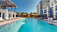 This outdoor amenity space is the centerpiece of a contemporary multi-story residential community, thoughtfully designed to deliver a resort-like experience. The space is anchored by a generously sized swimming pool with crystal-clear turquoise water, surrounded by an expansive pool deck finished in light-toned, large-format stone tiles that reflect the abundant natural sunlight. A dedicated shallow-entry area and perimeter safety fencing provide both accessibility and security throughout.

The pool deck is well-appointed with rows of white lounge chairs and cushioned chaise lounges, complemented by bold blue market umbrellas that offer shaded relaxation options. A built-in fireplace feature serves as a striking architectural focal point at the far end of the pool, framed by lush, mature palm trees that add a tropical ambiance to the setting. Additional seating and lounge areas are distributed across both sides of the deck, creating versatile spaces for gathering or quiet retreat.

The surrounding multi-story building features a modern façade with a mix of blue and white paneling, private balconies, and large windows that overlook the pool area. A covered pergola structure and an elevated terrace add architectural interest and additional outdoor living spaces. Gated entry points with wrought-iron fencing define the boundaries of the amenity space, while clean walkways and access corridors connect seamlessly to the broader community.