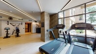 Located at 1 Fineran Way in Short Hills, NJ, this contemporary fitness center is thoughtfully designed to deliver a premium workout experience. The space is illuminated by sleek linear recessed lighting along the ceiling, complemented by abundant natural light streaming through floor-to-ceiling windows that overlook the surrounding exterior. Light wood-style flooring extends throughout, lending warmth and a clean aesthetic to the environment.

The facility is comprehensively equipped with a full suite of cardio machines, including treadmills, upright and recumbent bikes, and stair climbers, as well as a robust selection of strength training equipment such as cable machines, a multi-station functional trainer, adjustable benches, and a complete dumbbell rack. A dedicated open-floor zone provides space for functional training, featuring a wooden wall-mounted ladder, a BOSU ball, and other accessories.

Decorative geometric wall panels in warm neutral tones add architectural interest, while striking athlete-inspired murals enliven the walls, contributing to an energizing ambiance. A wall-mounted flat-screen TV and built-in storage cabinetry offer added convenience. The space is finished with proper ventilation and ceiling fans, ensuring comfort throughout any workout. Double glass doors provide easy access, completing this well-appointed fitness facility.