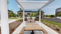 This outdoor communal space is designed to serve as a gathering and entertainment hub within a residential community. The centerpiece of the area is a striking white pergola supported by classic column posts, providing partial shade while maintaining an open, airy atmosphere that allows natural sunlight to filter through. Beneath the pergola, natural wood picnic tables with attached bench seating offer comfortable dining and socializing space for residents and their guests. At the rear of the pergola, two stainless steel gas grills are mounted on sturdy, built-in brick bases, providing a practical and attractive grilling station ideal for outdoor cooking and community events. The surrounding landscape is thoughtfully maintained, featuring manicured greenery, young trees, and decorative plantings that contribute to a welcoming environment. The space is set against a backdrop of multi-story residential buildings, emphasizing its role as a shared amenity within the broader community. The clean, well-kept paved surfaces and organized layout reflect a well-planned outdoor living area. Overall, the space strikes a balance between functionality and aesthetic appeal, making it an inviting destination for outdoor dining, grilling, and community gatherings throughout the warmer months.