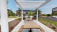 This outdoor communal space is designed to serve as a gathering and entertainment hub within a residential community. The centerpiece of the area is a striking white pergola supported by classic column posts, providing partial shade while maintaining an open, airy atmosphere that allows natural sunlight to filter through. Beneath the pergola, natural wood picnic tables with attached bench seating offer comfortable dining and socializing space for residents and their guests. At the rear of the pergola, two stainless steel gas grills are mounted on sturdy, built-in brick bases, providing a practical and attractive grilling station ideal for outdoor cooking and community events. The surrounding landscape is thoughtfully maintained, featuring manicured greenery, young trees, and decorative plantings that contribute to a welcoming environment. The space is set against a backdrop of multi-story residential buildings, emphasizing its role as a shared amenity within the broader community. The clean, well-kept paved surfaces and organized layout reflect a well-planned outdoor living area. Overall, the space strikes a balance between functionality and aesthetic appeal, making it an inviting destination for outdoor dining, grilling, and community gatherings throughout the warmer months.