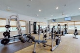 This fitness center is a thoughtfully designed, single-floor facility offering a wide range of equipment to support varied workout routines. The space is generously proportioned, allowing ample room to move freely between stations without feeling crowded.

The cardio zone lines one side of the room, featuring multiple treadmills, elliptical trainers, and a stationary bike, each positioned in front of a row of windows dressed with woven bamboo shades that admit generous natural light while maintaining a comfortable ambiance. Wall-mounted flat-screen TVs are positioned throughout the room, providing entertainment during workouts.

The strength training area is equally well-appointed, offering a multi-station cable machine, free weights including a full rack of dumbbells, barbells with weight plates, and adjustable benches. Large floor mirrors along one wall expand the sense of space and allow users to monitor form. Rubber flooring underlies the free weight section, while neutral-toned carpet covers the remainder of the room.

A small refreshment station with dark cabinetry, a quartz countertop, mosaic tile backsplash, and a built-in beverage cooler provides added convenience. Recessed lighting and ceiling fans complete the clean, functional design of this well-maintained facility.