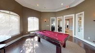 This single-story community clubhouse is thoughtfully designed to offer a variety of social and recreational spaces. The game room serves as a standout feature, centered around a full-size pool table with a bold red felt surface. The room is defined by its octagonal layout, warm taupe walls, elegant crown molding, and a series of arched windows that flood the space with natural light and offer views of the surrounding greenery. Wall sconces and recessed lighting add to the inviting atmosphere.

Adjacent to the game room is a comfortable lounge area, anchored by a generously sized sectional sofa and a rustic round coffee table. A striking orb-style chandelier serves as a focal point overhead, while large windows continue the theme of natural light throughout. A flat-screen television and media console make this a versatile space for both relaxing and entertaining.

The kitchen is fully equipped with rich wood cabinetry, light stone countertops, stainless steel appliances including a full-size refrigerator and range, and large-format tile flooring. Crown molding and a window above the counter tie the space together with a polished, cohesive finish. Together, these spaces create a functional and welcoming environment ideal for gatherings and community use.