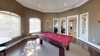 This single-story community clubhouse is thoughtfully designed to offer a variety of social and recreational spaces. The game room serves as a standout feature, centered around a full-size pool table with a bold red felt surface. The room is defined by its octagonal layout, warm taupe walls, elegant crown molding, and a series of arched windows that flood the space with natural light and offer views of the surrounding greenery. Wall sconces and recessed lighting add to the inviting atmosphere.

Adjacent to the game room is a comfortable lounge area, anchored by a generously sized sectional sofa and a rustic round coffee table. A striking orb-style chandelier serves as a focal point overhead, while large windows continue the theme of natural light throughout. A flat-screen television and media console make this a versatile space for both relaxing and entertaining.

The kitchen is fully equipped with rich wood cabinetry, light stone countertops, stainless steel appliances including a full-size refrigerator and range, and large-format tile flooring. Crown molding and a window above the counter tie the space together with a polished, cohesive finish. Together, these spaces create a functional and welcoming environment ideal for gatherings and community use.