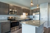 This single-floor unit offers a thoughtfully designed open-concept layout that seamlessly connects the kitchen and living area. The kitchen is a standout feature, showcasing rich dark-toned shaker cabinetry, light granite countertops with subtle veining, and a large center island ideal for dining or meal prep. Stainless steel appliances — including a French door refrigerator, electric range, built-in microwave, and dishwasher — complete the space, while elegant gold pendant lighting adds a contemporary touch. The living area extends naturally from the kitchen, featuring hardwood-style flooring, a ceiling fan, and a sliding glass door that floods the space with natural light and provides outdoor views. Bedrooms are well-proportioned, each with hardwood-style flooring, ceiling fans, and ample natural light from large windows with blinds. One bedroom includes access to a spacious walk-in closet with extensive shelving and rod storage, connecting to a dedicated bathroom. The bathroom features matching dark cabinetry, a granite vanity top, tile flooring, and a well-lit mirror with contemporary fixtures. Throughout the unit, neutral gray walls and clean white trim create a cohesive, modern aesthetic.