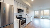 Located at 7900 SE Luther Rd in Portland, Oregon, this single-floor unit offers a clean, modern aesthetic with thoughtfully selected finishes throughout. The open-concept living and kitchen area is bright and welcoming, with light hardwood flooring that runs continuously through the space. Large windows and a sliding glass door flood the living area with natural light and provide views of the outdoor surroundings, including a landscaped area with an iron railing fence.

The kitchen is well-appointed with white shaker-style cabinetry, open shelving, and a striking geometric hexagonal tile backsplash. Stainless steel appliances — including a refrigerator, electric range, over-the-range microwave, and dishwasher — complement the clean white countertops and matte black hardware and fixtures, delivering a contemporary, functional workspace.

The bathroom features navy blue shaker-style cabinetry with a white countertop, an undermount sink, a large vanity mirror with stylish globe lighting, and a full tub-shower combination. The bedroom includes a dedicated walk-in closet, offering practical storage.

A stacked washer and dryer unit is conveniently integrated within the unit. The overall design is cohesive and modern, making effective use of the available space while maintaining a bright, airy atmosphere throughout.